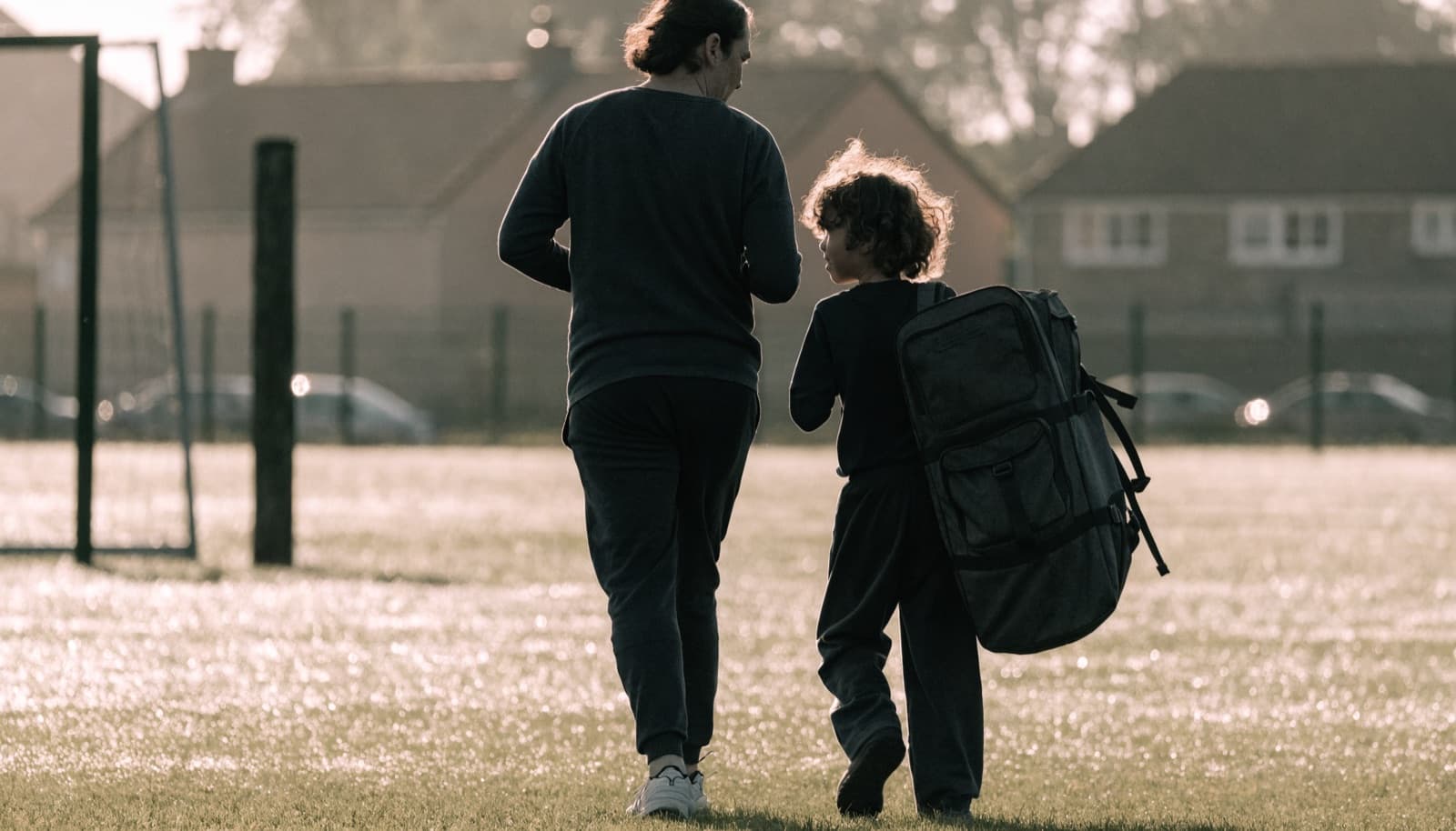 A parent and child walk to morning training across a dewy pitch, the child carrying a kit bag almost as big as them